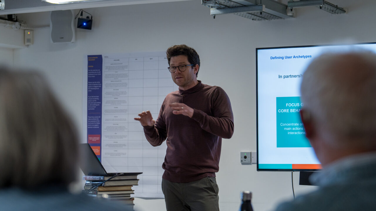 A man giving a presentation in a meeting room. He is standing near a large screen displaying a slide titled 'Defining User Archetypes'. The slide includes a section that reads 'FOCUS ON CORE BEHAVIOURS'. The presenter is speaking with hand gestures, and several people are seated in the foreground, listening. A laptop rests on a stack of books on the table beside him. Dyn yn rhoi cyflwyniad mewn ystafell gyfarfod. Mae'n sefyll ger sgrin fawr sy'n arddangos sleid o'r enw 'Diffinio Archeteipiau Defnyddwyr'. Mae'r sleid yn cynnwys adran sy'n darllen 'FFOCWS AR YMDDANGOSIADAU CRAIDD'. Mae'r cyflwynydd yn siarad â symudiadau llaw, ac mae sawl person yn eistedd yn y blaendir, yn gwrando. Mae gliniadur yn gorffwys ar bentwr o lyfrau ar y bwrdd wrth ei ymyl.