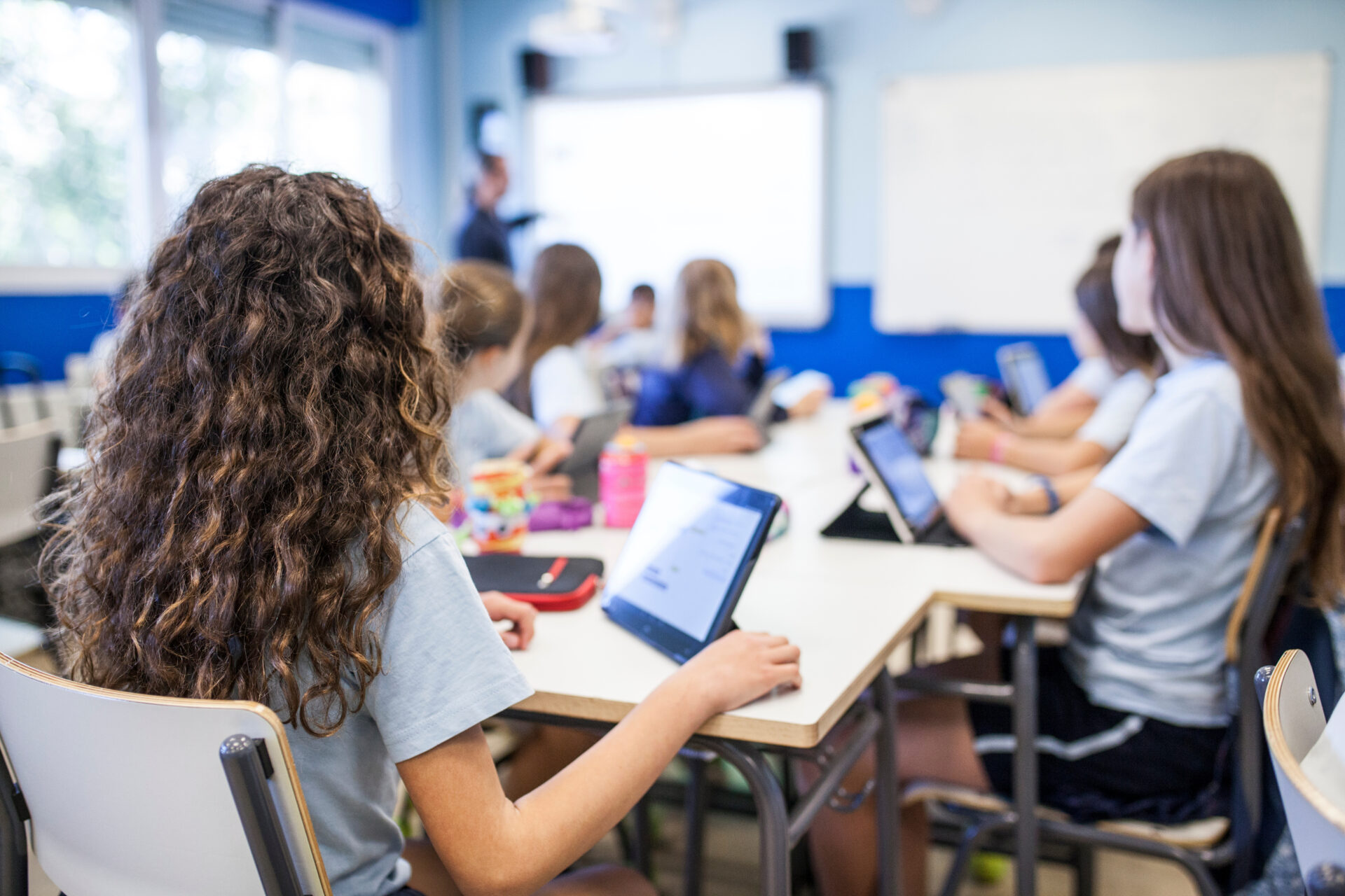 girl with brown curly hair studies in class with her tablet next to her classmates while they listen to the teacher Merch gyda gwallt cyrliog brown yn astudio yn y dosbarth gyda’i thabled wrth ochr ei chyfoedion tra maen nhw’n gwrando ar y tiwtor.