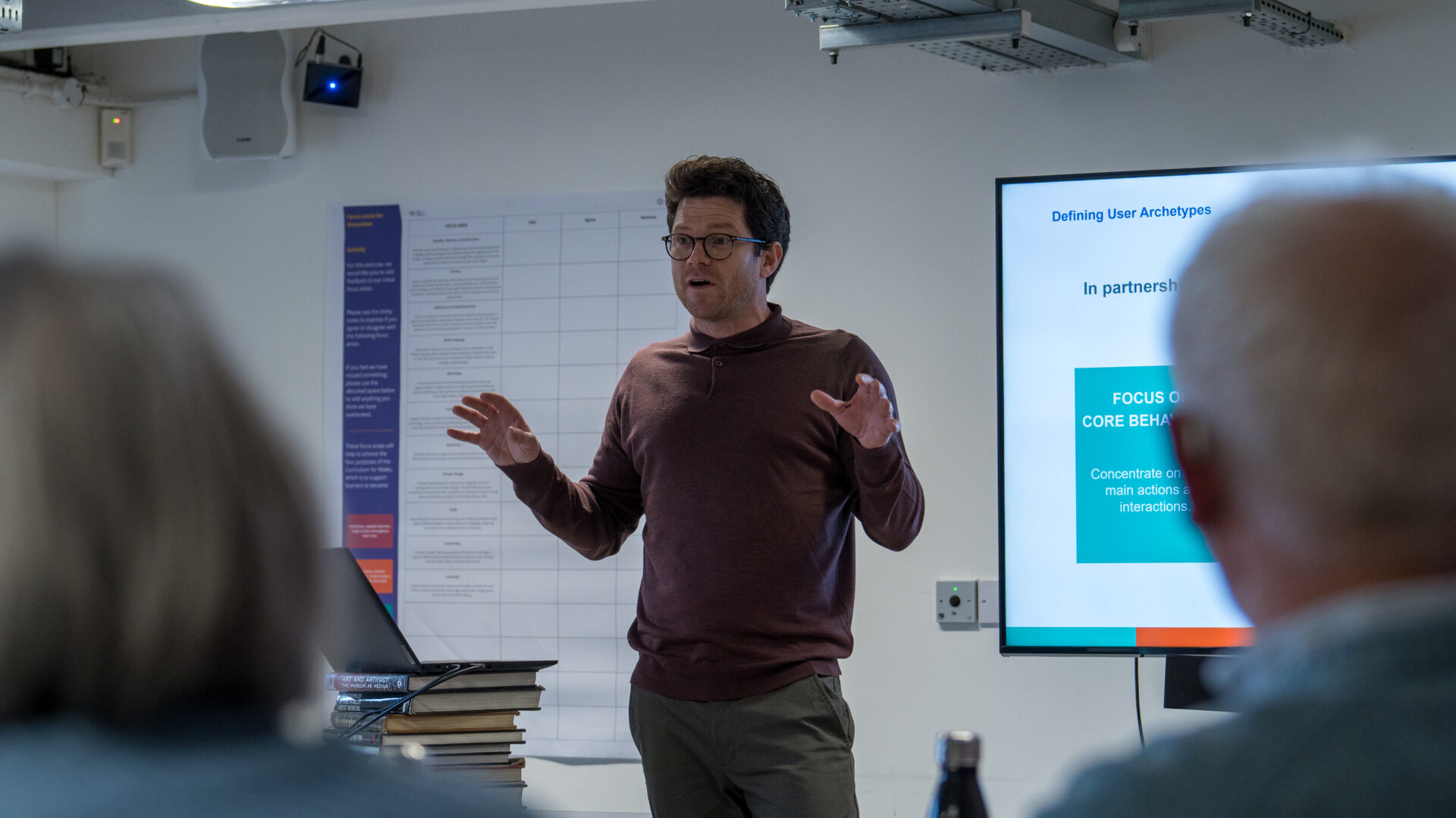 A man gives a presentation to a seated audience, speaking in front of a screen and a wall chart. Dyn yn rhoi cyflwyniad i gynulleidfa eisteddedig, yn siarad o flaen sgrin a siart ar y wal.
