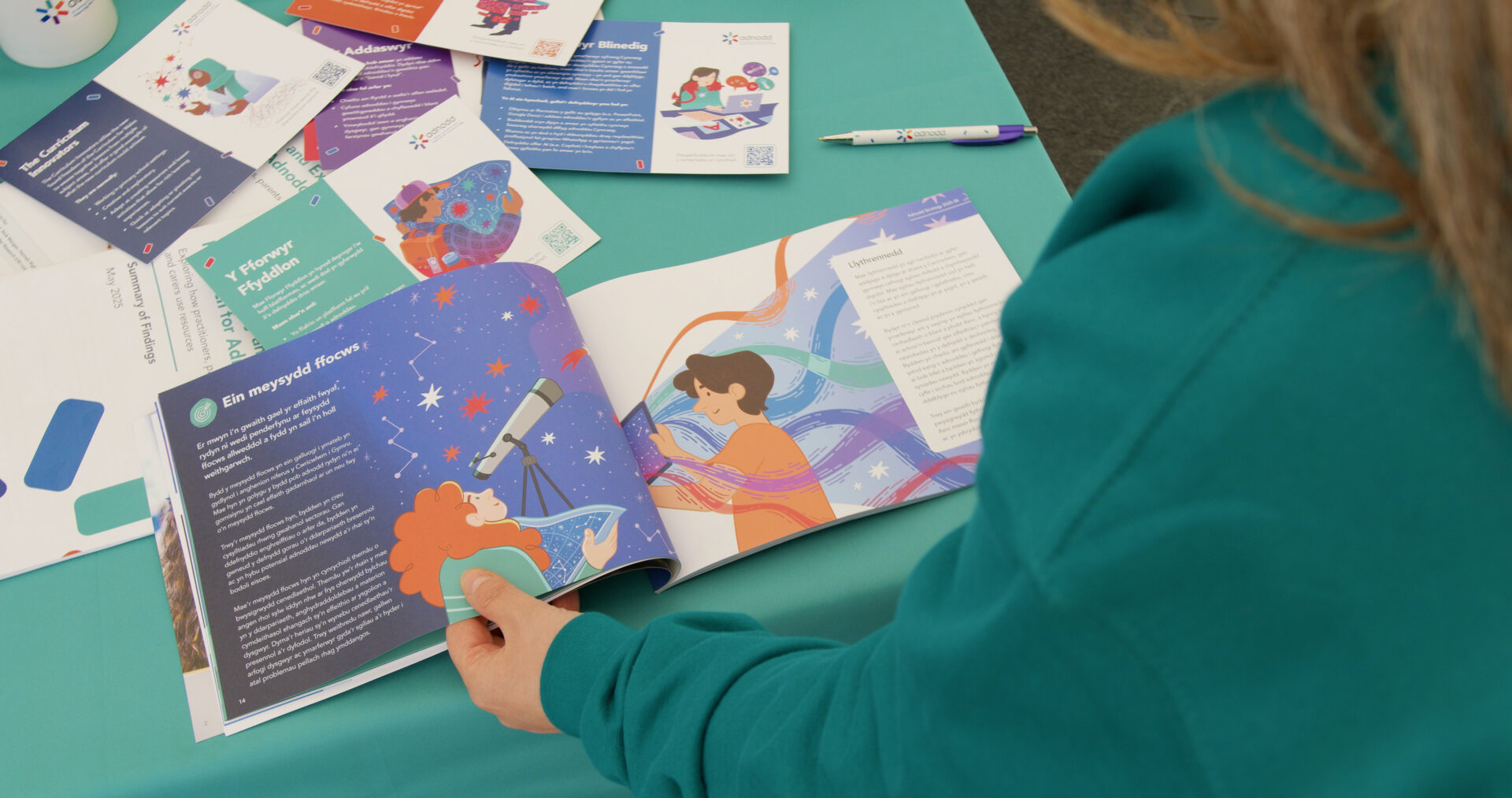 A person in a teal jumper is reading a colourful booklet at a display table covered with leaflets and resources from Adnodd. Mae person mewn siwmper laswyrdd yn darllen llyfryn lliwgar wrth ddesg sy'n llawn taflenni a deunyddiau gan Adnodd.