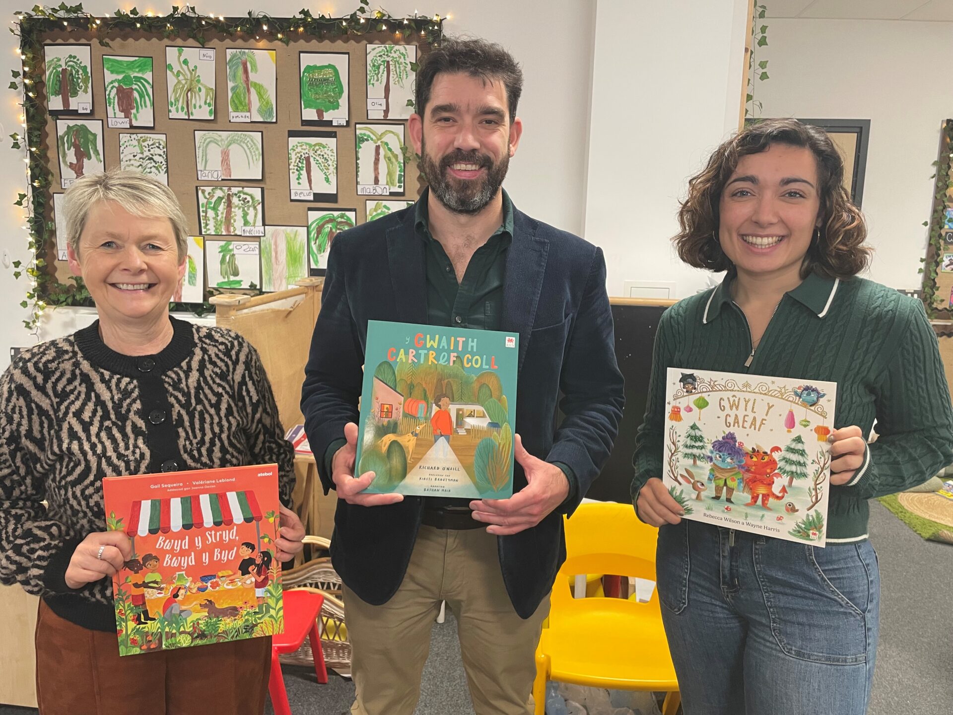 Three smiling adults standing in a classroom, each holding a colourful children’s book in Welsh